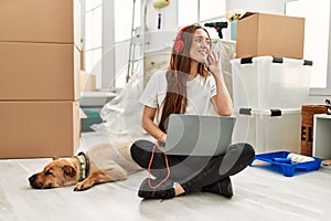 Young hispanic woman listening to music sitting on floor with dog at new home
