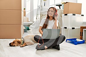 Young hispanic woman listening to music sitting on floor with dog at new home