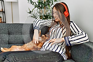 Young hispanic woman listening to music sitting on sofa with dog at home