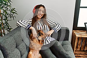 Young hispanic woman listening to music sitting on sofa with dog at home