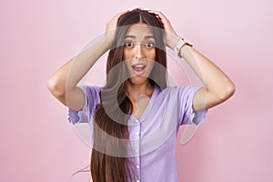 Young hispanic woman with long hair standing over pink background crazy and scared with hands on head, afraid and surprised of