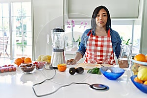 Young hispanic woman making healthy smoothie with serious expression on face