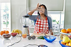 Young hispanic woman making healthy smoothie smiling happy doing ok sign with hand on eye looking through fingers
