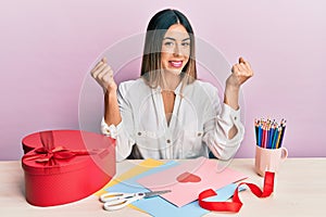 Young hispanic woman making valentine gift sitting on the table very happy and excited doing winner gesture with arms raised,