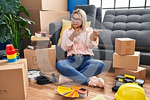 Young hispanic woman moving to a new home sitting on the floor pointing fingers to camera with happy and funny face