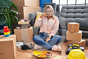 Young hispanic woman moving to a new home sitting on the floor smiling doing phone gesture with hand and fingers like talking on