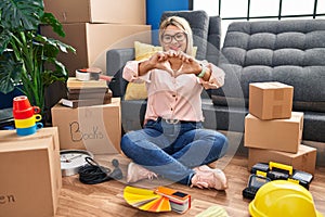 Young hispanic woman moving to a new home sitting on the floor smiling in love doing heart symbol shape with hands