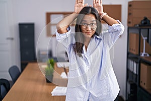 Young hispanic woman at the office doing bunny ears gesture with hands palms looking cynical and skeptical