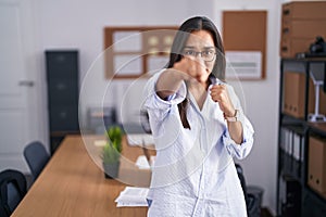 Young hispanic woman at the office punching fist to fight, aggressive and angry attack, threat and violence