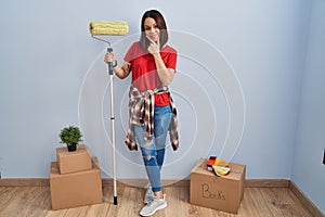 Young hispanic woman painting home walls with paint roller looking confident at the camera smiling with crossed arms and hand