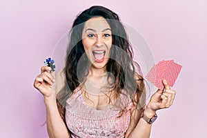 Young hispanic woman playing poker holding casino chips and cards celebrating crazy and amazed for success with open eyes