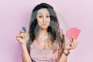 Young hispanic woman playing poker holding casino chips and cards puffing cheeks with funny face