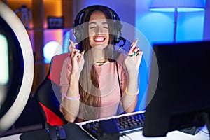 Young hispanic woman playing video games gesturing finger crossed smiling with hope and eyes closed