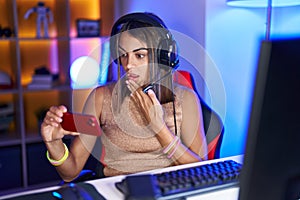Young hispanic woman playing video games with smartphone covering mouth with hand, shocked and afraid for mistake
