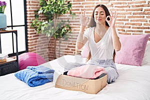 Young hispanic woman putting used clothes into donations box sitting on the bed relax and smiling with eyes closed doing