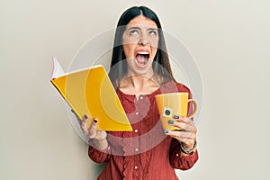 Young hispanic woman reading book drinking cup of tea angry and mad screaming frustrated and furious, shouting with anger looking