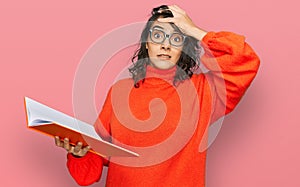 Young hispanic woman reading a book wearing glasses stressed and frustrated with hand on head, surprised and angry face