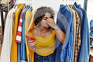 Young hispanic woman searching clothes on clothing rack using smartphone tired rubbing nose and eyes feeling fatigue and headache