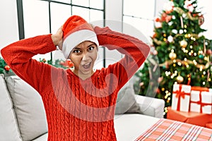 Young hispanic woman with short hair wearing christmas hat sitting on the sofa crazy and scared with hands on head, afraid and