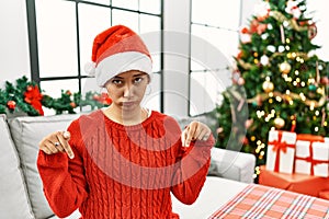 Young hispanic woman with short hair wearing christmas hat sitting on the sofa pointing down looking sad and upset, indicating
