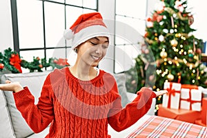 Young hispanic woman with short hair wearing christmas hat sitting on the sofa smiling showing both hands open palms, presenting