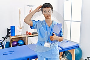 Young hispanic woman with short hair working at pain recovery clinic confuse and wondering about question