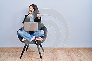 Young hispanic woman sitting on chair using computer laptop pointing fingers to camera with happy and funny face