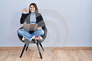 Young hispanic woman sitting on chair using computer laptop smiling positive doing ok sign with hand and fingers