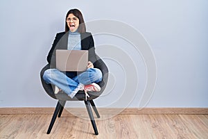 Young hispanic woman sitting on chair using computer laptop sticking tongue out happy with funny expression