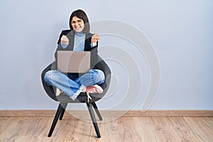Young hispanic woman sitting on chair using computer laptop very happy and excited doing winner gesture with arms raised, smiling