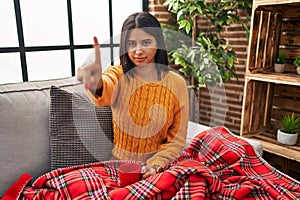 Young hispanic woman sitting on the sofa drinking a coffee at home pointing with finger up and angry expression, showing no