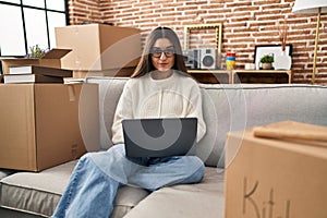 Young hispanic woman sitting on the sofa at new home using laptop happy face smiling with crossed arms looking at the camera