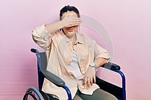 Young hispanic woman sitting on wheelchair covering eyes with hand, looking serious and sad