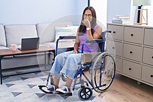 Young hispanic woman sitting on wheelchair at home smelling something stinky and disgusting, intolerable smell, holding breath