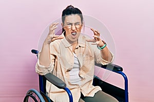 Young hispanic woman sitting on wheelchair shouting frustrated with rage, hands trying to strangle, yelling mad