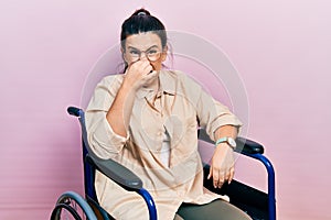 Young hispanic woman sitting on wheelchair smelling something stinky and disgusting, intolerable smell, holding breath with