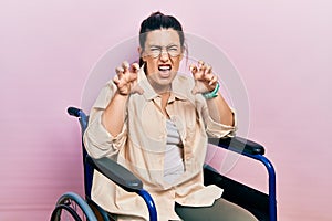 Young hispanic woman sitting on wheelchair smiling funny doing claw gesture as cat, aggressive and sexy expression