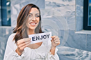 Young hispanic woman smiling happy holding enjoy word paper standing at the city