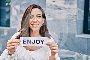 Young hispanic woman smiling happy holding enjoy word paper standing at the city