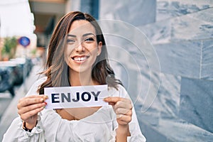 Young hispanic woman smiling happy holding enjoy word paper standing at the city