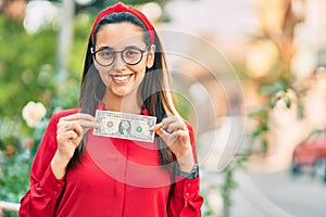 Young hispanic woman smiling happy holding one usa dollar banknote at the city