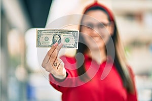 Young hispanic woman smiling happy holding one usa dollar banknote at the city