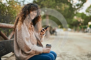 Young hispanic woman smiling happy using smartphone and drinking coffee at the park