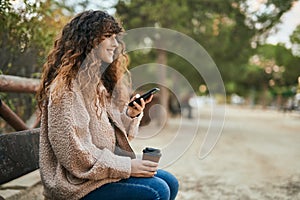 Young hispanic woman smiling happy using smartphone and drinking coffee at the park