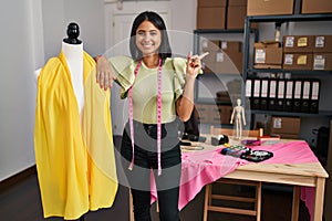 Young hispanic woman standing by manikin at small business smiling happy pointing with hand and finger to the side