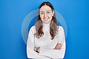 Young hispanic woman standing over blue background happy face smiling with crossed arms looking at the camera