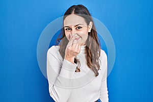Young hispanic woman standing over blue background smelling something stinky and disgusting, intolerable smell, holding breath