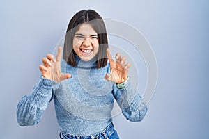 Young hispanic woman standing over blue background smiling funny doing claw gesture as cat, aggressive and sexy expression