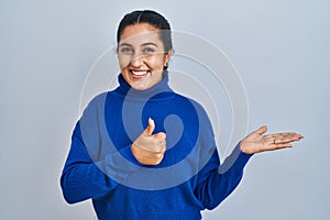 Young hispanic woman standing over isolated background showing palm hand and doing ok gesture with thumbs up, smiling happy and