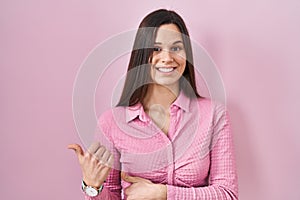 Young hispanic woman standing over pink background pointing to the back behind with hand and thumbs up, smiling confident